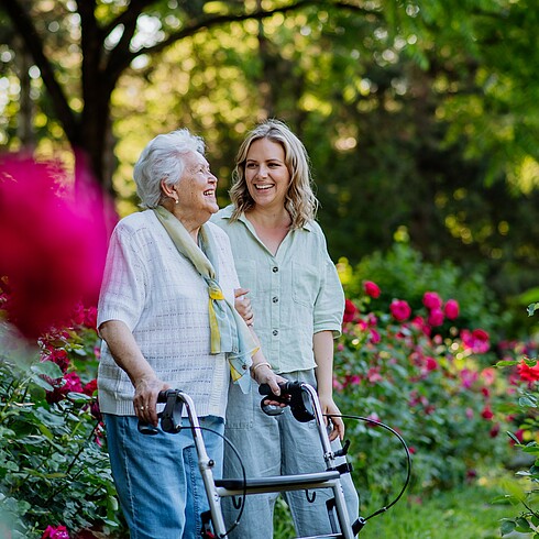 Eine ältere Frau geht auf einen Rollator gestützt neben einer jüngeren Frau durch einen blühenden Garten.